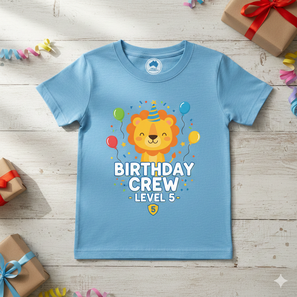 A light blue custom-printed kids' birthday t-shirt featuring a cute lion in a party hat and "Birthday Crew Level 5" text, displayed on a wooden background with party decorations.